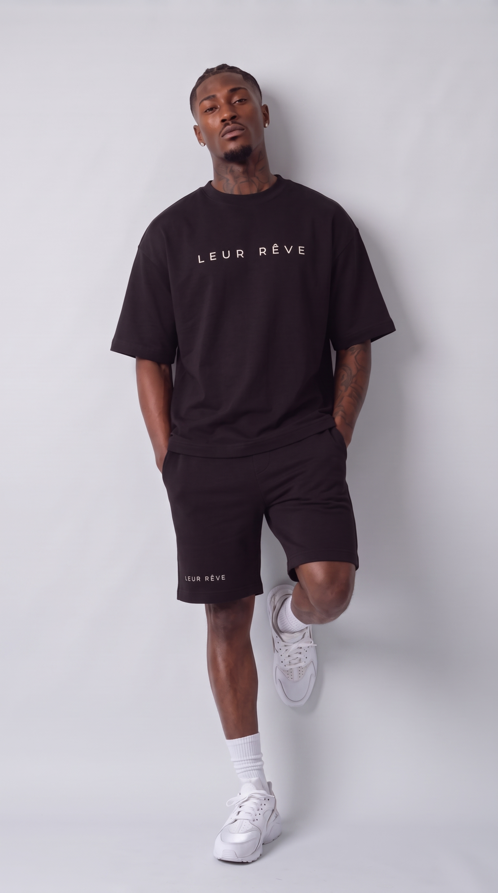Man wearing the Leur Rêve Short in black, leaning against the wall with one knee raised, wordmark at the hem