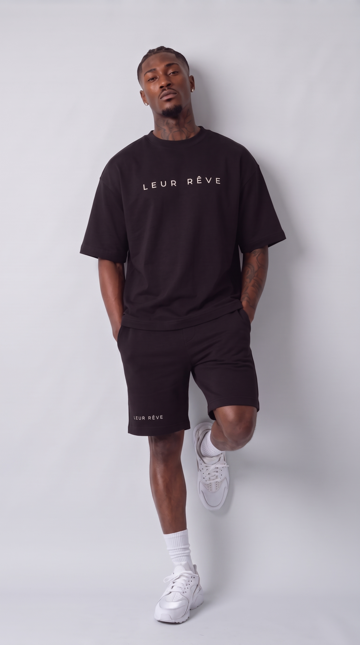 Man wearing the Leur Rêve Short in black, leaning against the wall with one knee raised, wordmark at the hem